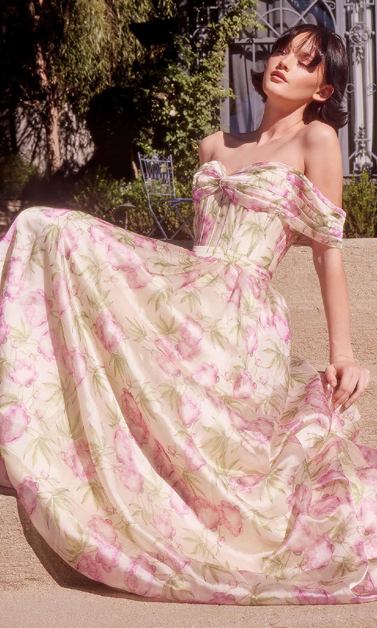 A woman sits outdoors in the Andrea & Leo A1466 long floral-print prom dress, its off-the-shoulder design, light pink and green blooms, and corset-style bodice beautifully highlighted by the sunlight.